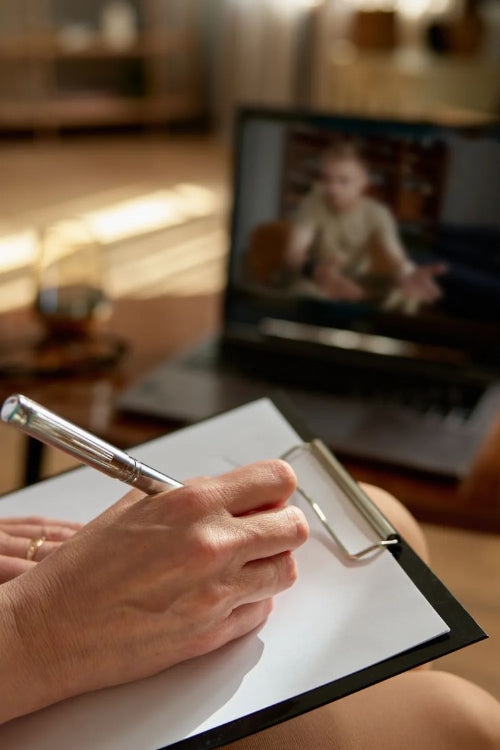 Person taking notes with a pen on an NDIS Functional capacitity report (FCA_) assessment form on a clipboard, with a support coordinator and participant in the background reviewing an NDIS plan on a laptop (soft focus), in a bright home or clinic setting.